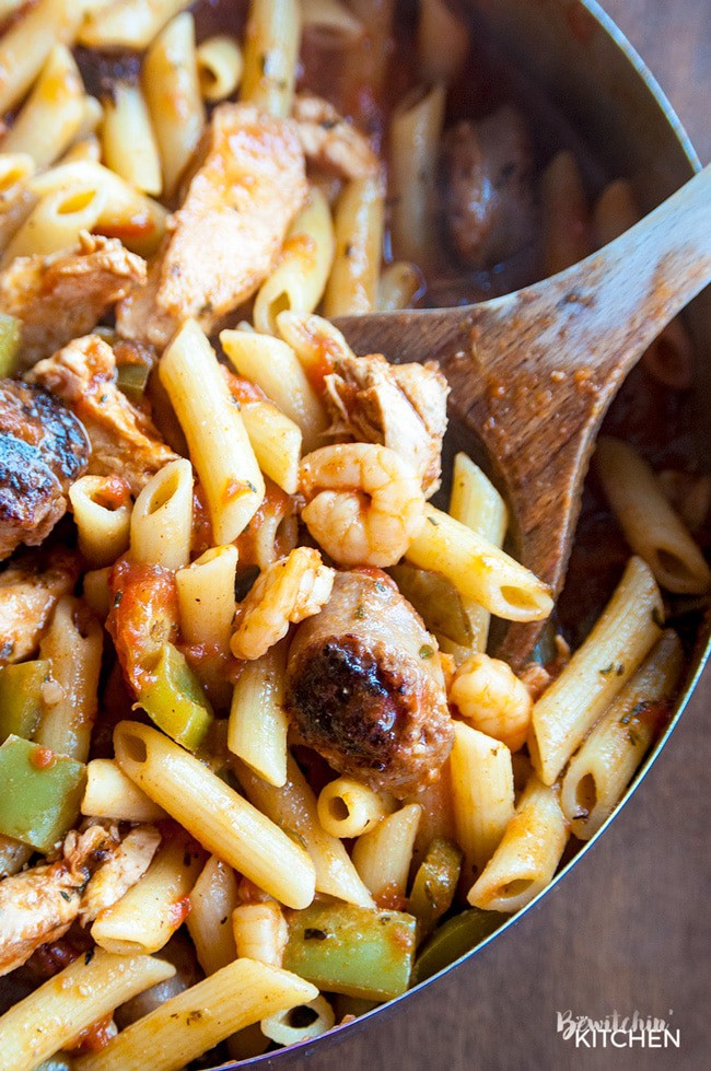 A wood spoon lifting jambalaya made with penne noodles out of a chef pan.