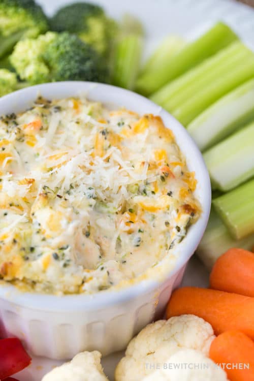 Hot broccoli dip with shaved parmesan on top surrounded by broccoli, celery, and carrots.