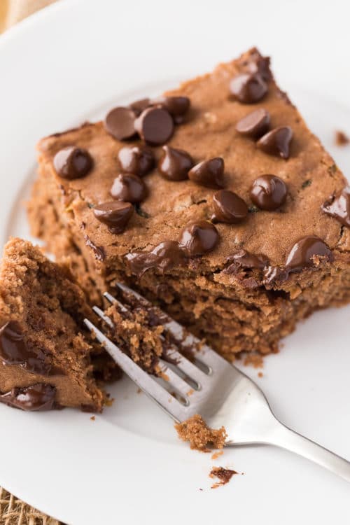 A piece of chocolate zucchini cake on a white plate with a fork cutting through the cake.