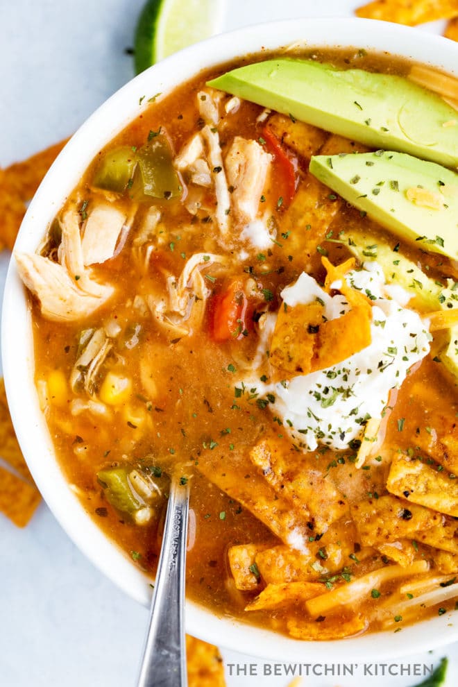 A bowl of chicken tortilla soup with avocado, greek yogurt, and tortilla strips. Sprinkled with tex mex cheese.