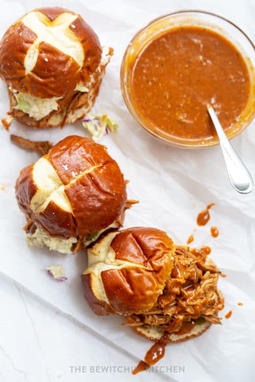 Looking down on three pulled pork sandwiches on a pretzel bun with a small bowl of homemade bbq sauce beside it.