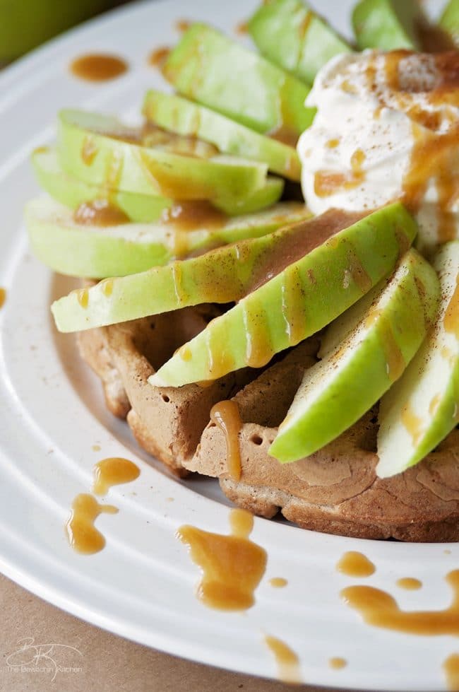 Side shot of a crispy whole wheat waffle topped with granny smith apples, caramel drizzle, and whipped cream.