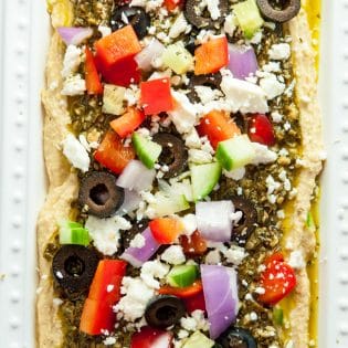 Layered Pesto Greek Dip - EASIEST DIP RECIPE EVER! This Mediterranean inspired appetizer uses hummus, pesto, feta, peppers, olives, and cucumbers. Goes great with veggies and chips. 21 Day Fix approved and a healthy appetizer recipe.