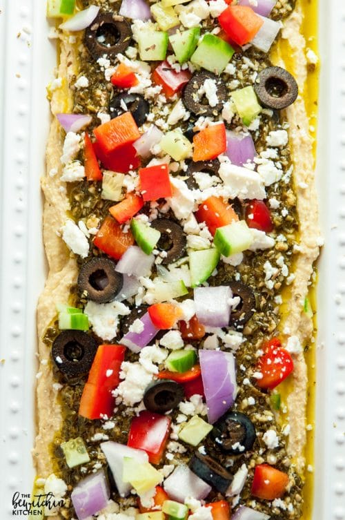 Layered Pesto Greek Dip - EASIEST DIP RECIPE EVER! This Mediterranean inspired appetizer uses hummus, pesto, feta, peppers, olives, and cucumbers. Goes great with veggies and chips. 21 Day Fix approved and a healthy appetizer recipe.