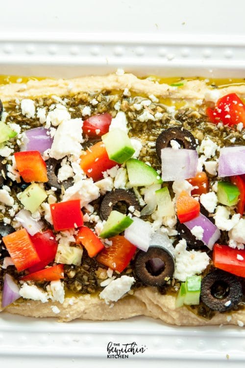 Layered Pesto Greek Dip - EASIEST DIP RECIPE EVER! This Mediterranean inspired appetizer uses hummus, pesto, feta, peppers, olives, and cucumbers. Goes great with veggies and chips. 21 Day Fix approved and a healthy appetizer recipe.