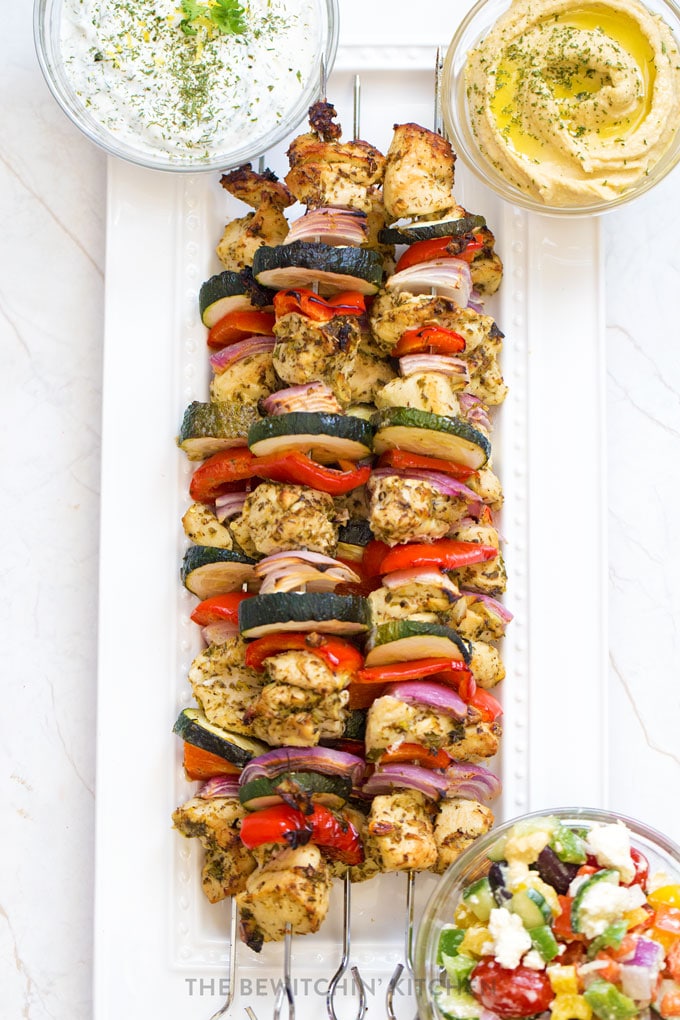 Chicken veggie kebabs on a white platter with hummus, tzatziki, and greek salad.