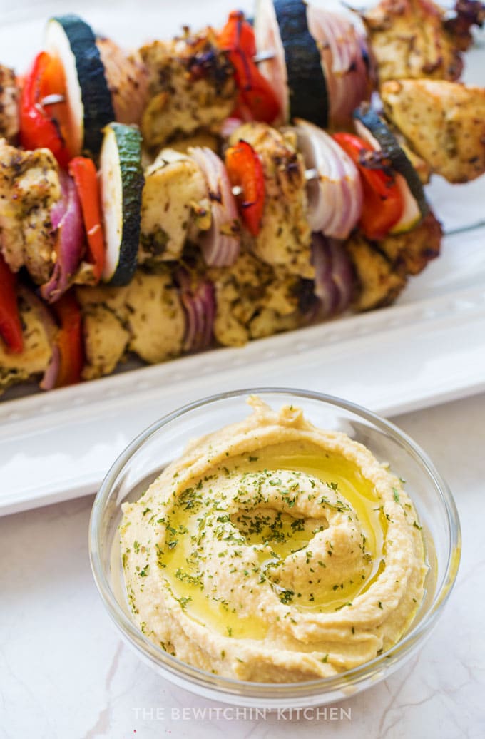 A bowl of hummus swirled with olive oil in the foreground with chicken souvlaki in the background.
