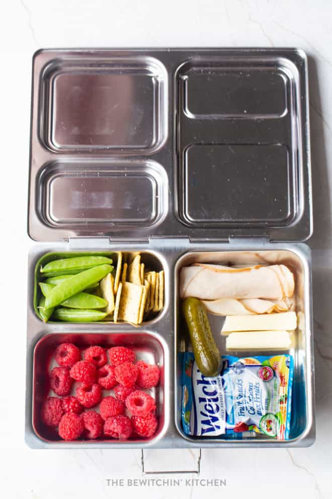 Snap peas, chickpea crackers, pickle, turkey roll up, string cheese, Welch's Fruit Snacks, and raspberries in a healthy bento box lunch.