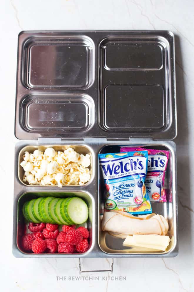 Simple and healthy school lunch idea - popcorn, sliced cucumbers, raspberries, Welch's Fruit Snacks, turkey roll ups, and string cheese in a Planetbox bento box for school lunch.