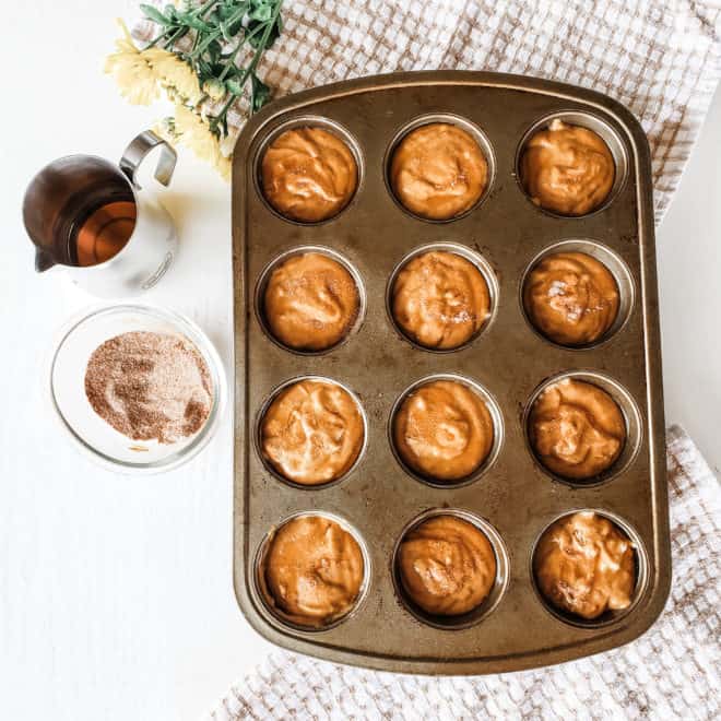 pumpkin muffin batter in tin surrounded by maple syrup and spiced topping