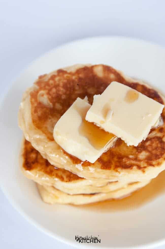 A stack of three gluten free buttermilk pancakes with two pats of butter and maple syrup on a white plate.