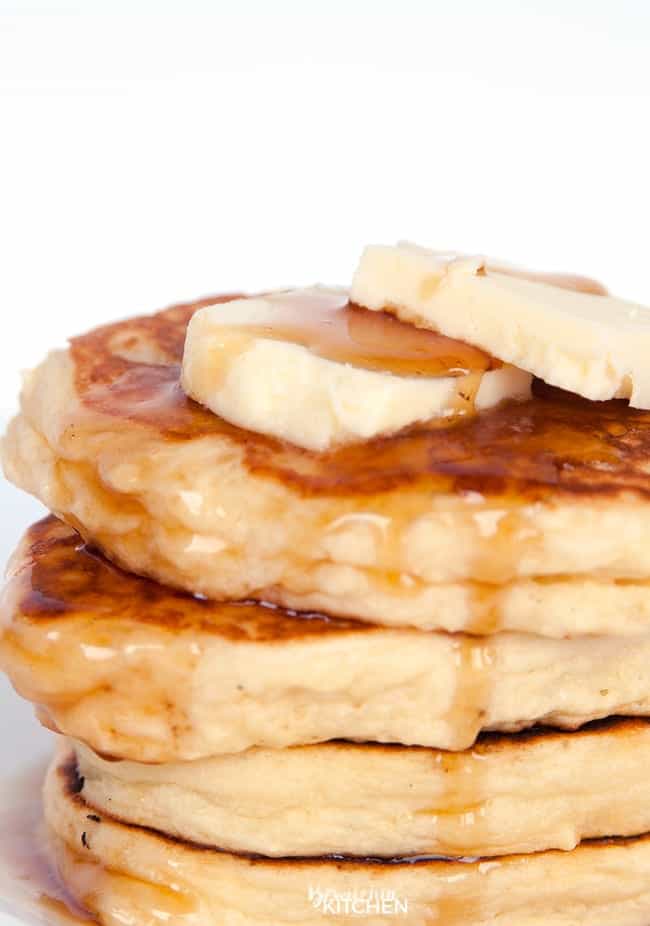 A stack of four gluten free buttermilk pancakes with two pats of butter and maple syrup on a white plate.
