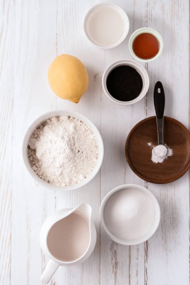 An aerial view of lemon poppy seed cake ingredients including five small white ramekins filled with lemon juice, oil, vanilla extract, baking soda, flour, and sugar, as well as a small white pitcher full of milk and one lemon.