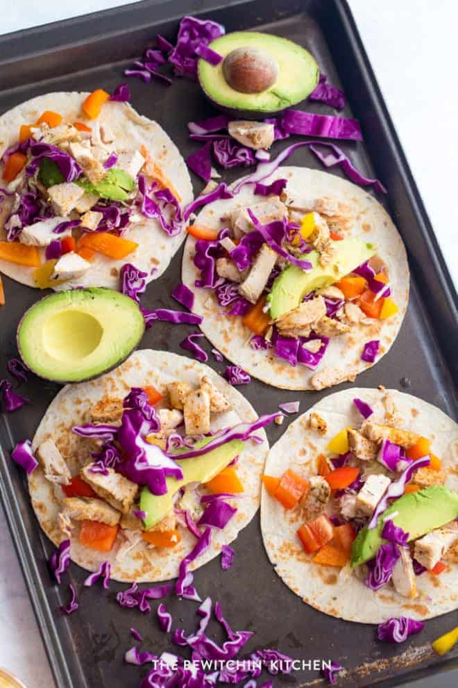 Top view of 4 grilled chicken tacos on a sheet pan with halves of avocados