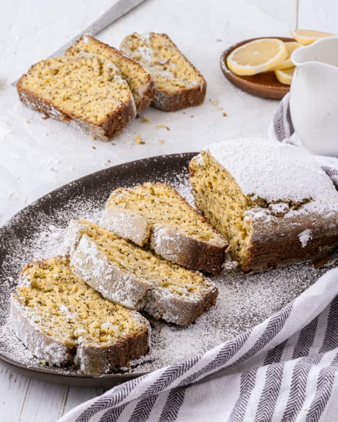 A lemon poppy seed pound cake with powdered sugar on top that has been sliced into four pieces on a dark brown plate. There are three other cut slices visible in the background, as well as a plate with sliced lemons and a small white pitcher. 