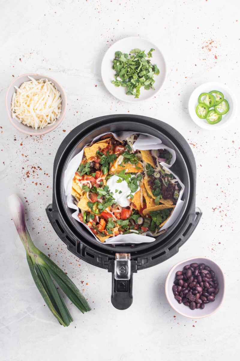Overhead of nachos in air fryer with bowls of ingredients scattered around the air fryer